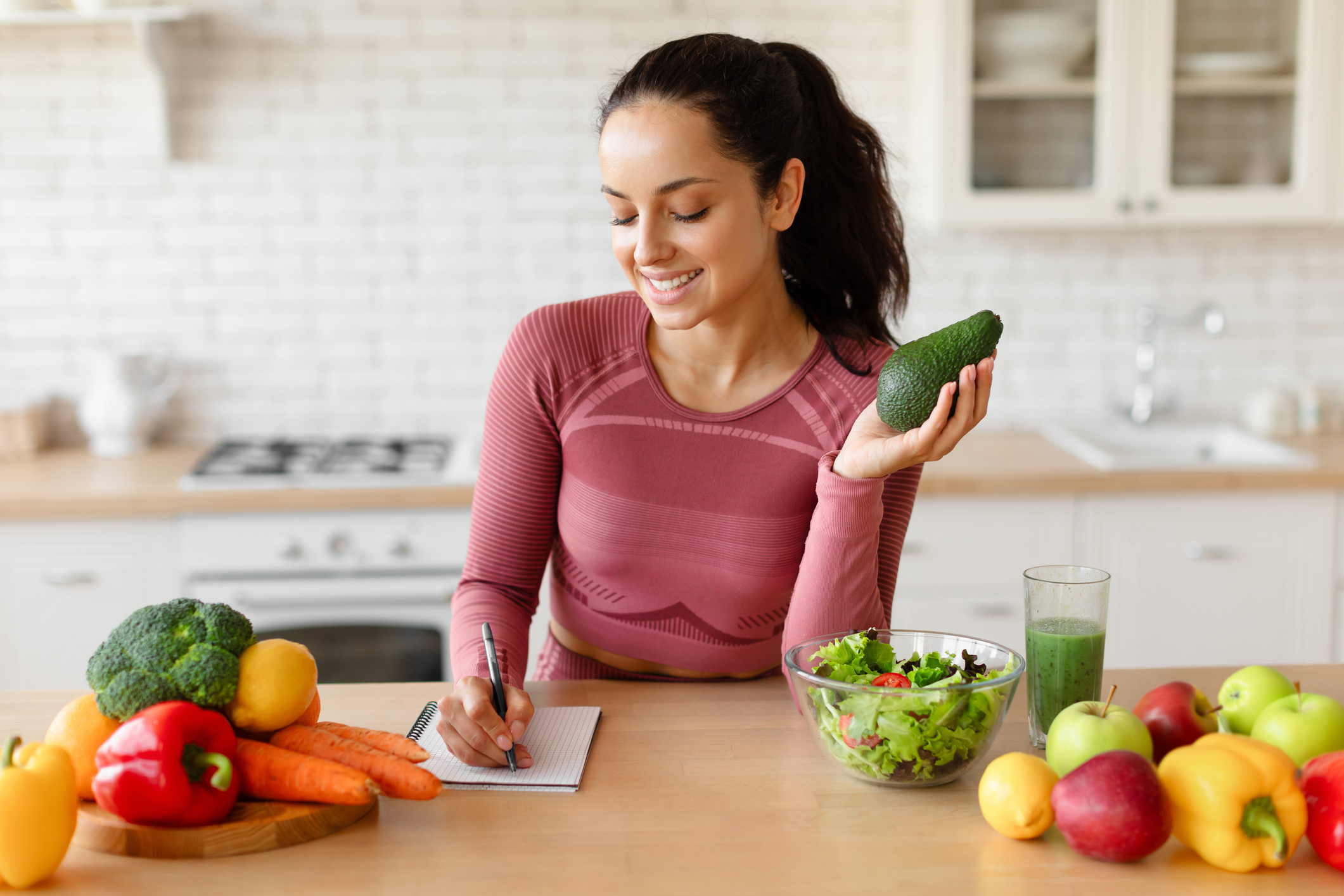 A woman holding a cucumber and writing recipes in the kitchen.