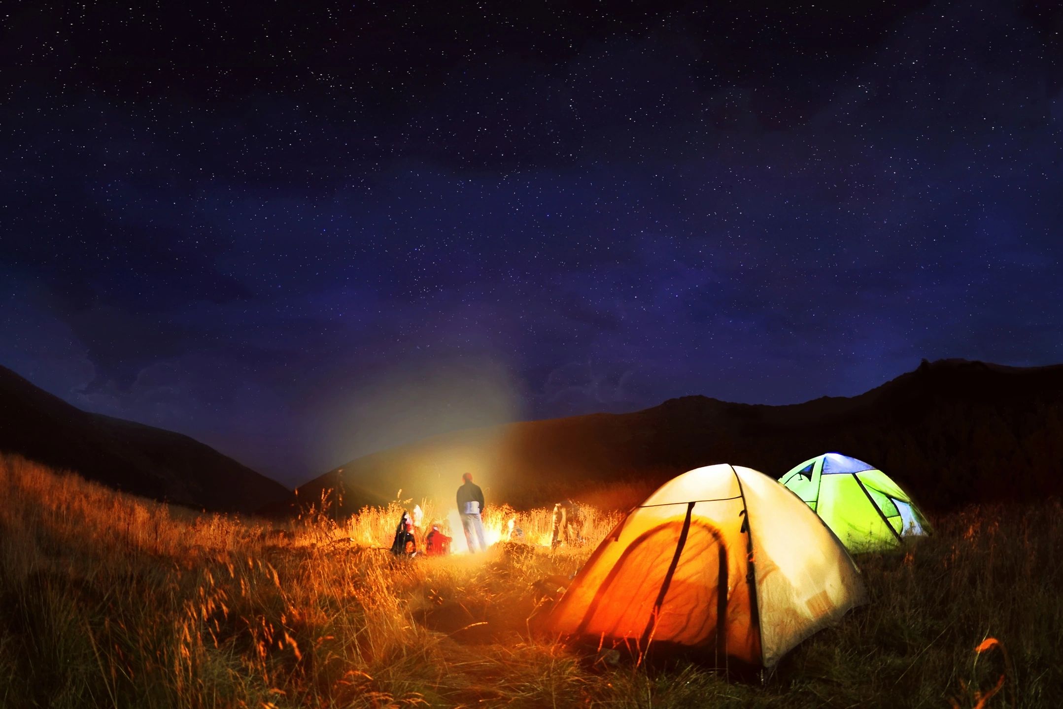 Campers gather around a campfire under a starry night sky near illuminated tents.