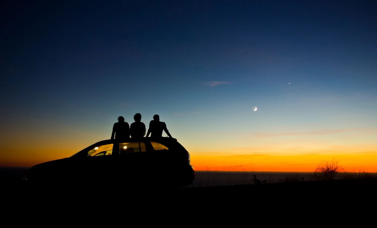 Three friends sitting on a car roof at sunset with a crescent moon.